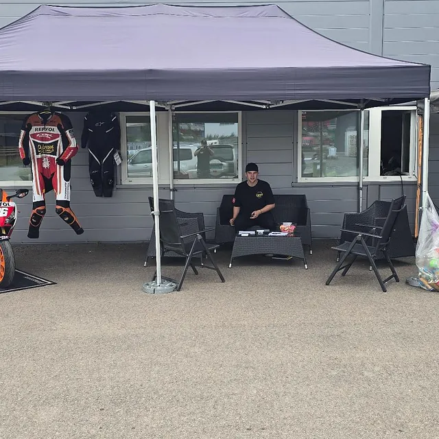 To telt utenfor et bygg med utstilt Repsol-motorsykkel og racingdress, samt en mann som sitter under tak.