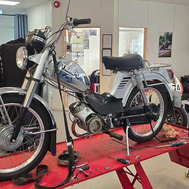En eldre Tempo lett motorsykkel står på et rødt løftebord i et verksted omgitt av verktøy og deler.